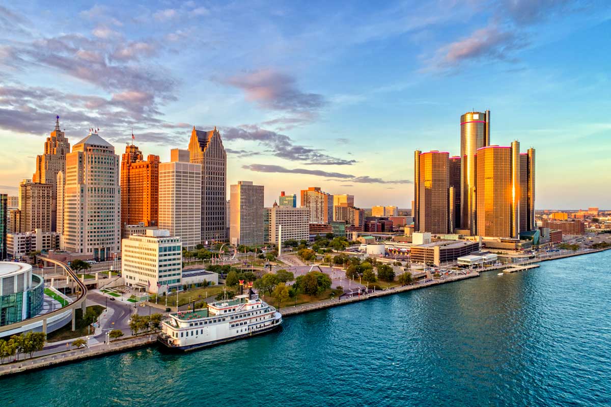 Aerial view of downtown Detroit, Michigan at sunset, showing modern skyscrapers, historic buildings, and the Detroit River with a white boat docked along the waterfront.