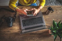 Person in a yellow sweater using a smartphone above a laptop on a wooden table. Nearby are a camera, a pair of glasses, a mug, and a potted plant. The setting suggests a workspace or home office environment.