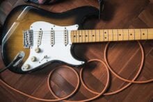 A close-up of an electric guitar with a sunburst finish and white pickguard lying on a wooden floor, with an orange cable coiled around it.