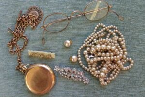A collection of vintage jewelry and accessories on a green fabric, including a gold pocket watch, eyeglasses, pearl necklaces, a jeweled brooch, pearl earrings, a decorative pin, and a chain.