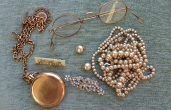 A collection of vintage jewelry and accessories on a green fabric, including a gold pocket watch, eyeglasses, pearl necklaces, a jeweled brooch, pearl earrings, a decorative pin, and a chain.