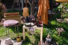 A garage sale setup outdoors displays a table with a "Garage Sale" sign, assorted dishes, a brown bag, a guitar, a yellow dress, a crochet cushion, a hat, a lamp, a small plant, and various household items on the grass.