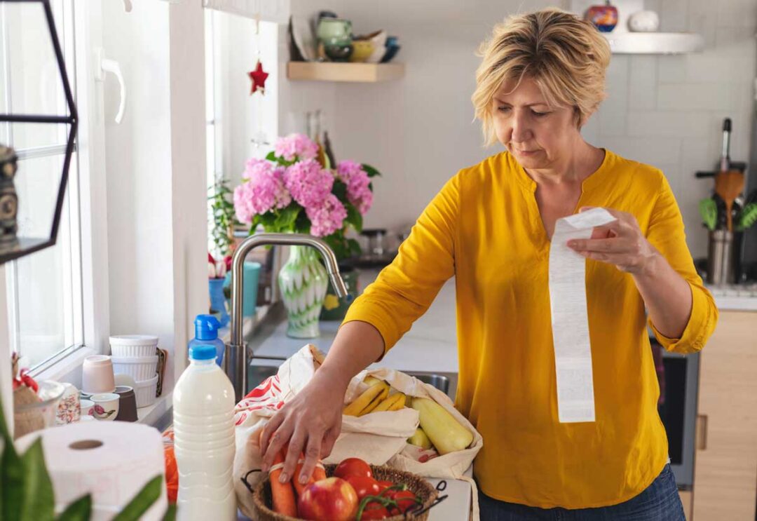 A woman in a yellow shirt stands in a kitchen, checking a long receipt while unpacking groceries, including fruits, vegetables, and a bottle of milk, with flowers visible on the counter in the background.