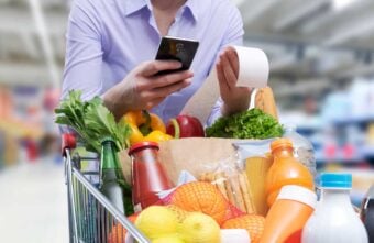 A person stands in a grocery store holding a receipt and a smartphone, with a shopping cart full of fresh produce, bread, bottled drinks, and packaged foods in the foreground.