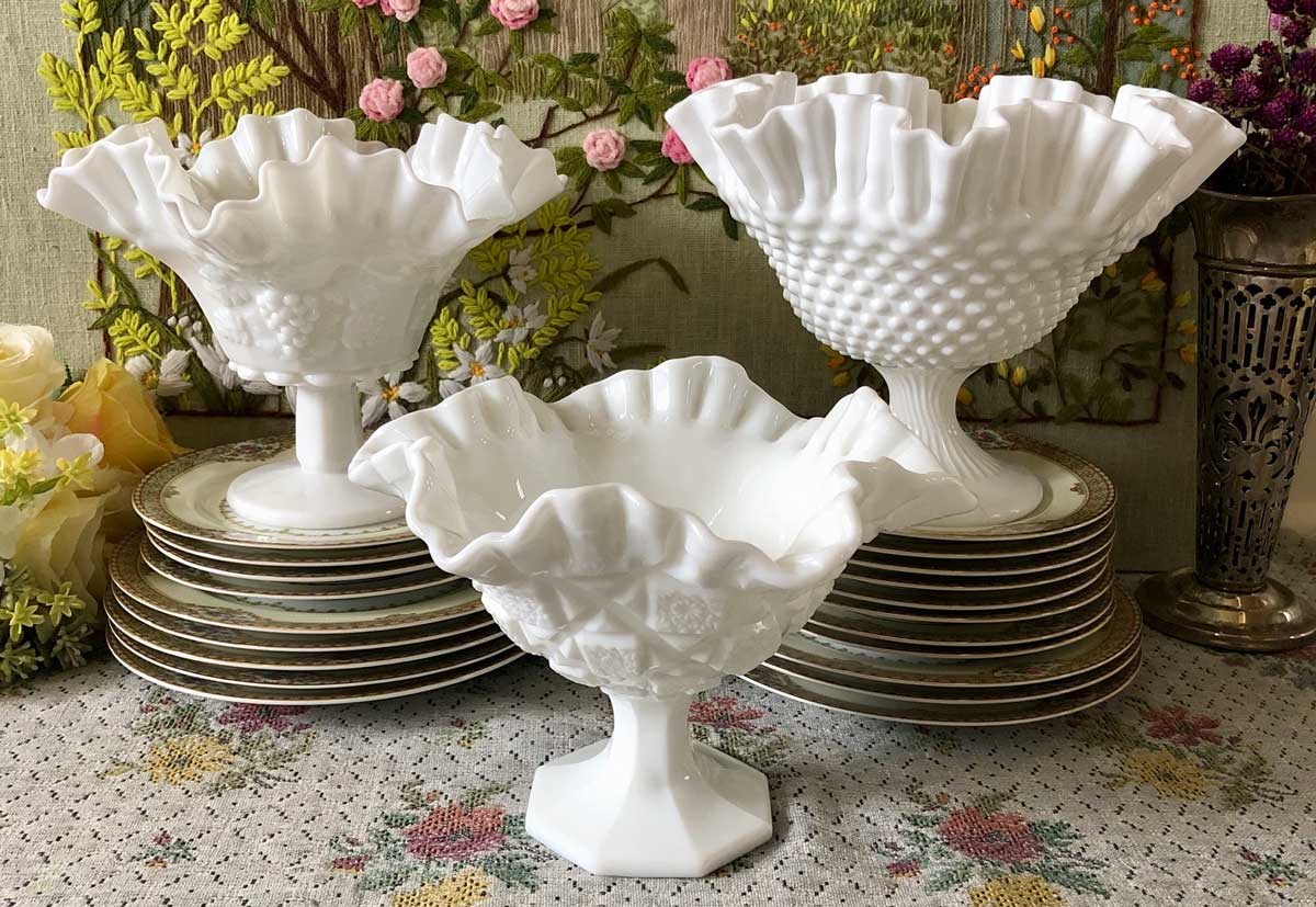 Three white milk glass pedestal bowls with ruffled edges are displayed in front of stacked floral plates on a table with a floral tablecloth. Flowers and greenery are in the background.