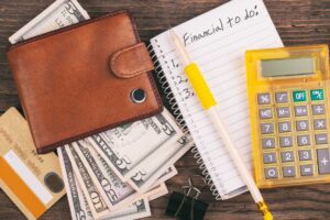 A brown wallet with cash, credit cards, a yellow calculator, a pencil, and a notepad labeled “Financial to do” are arranged on a wooden desk, suggesting personal finance planning.