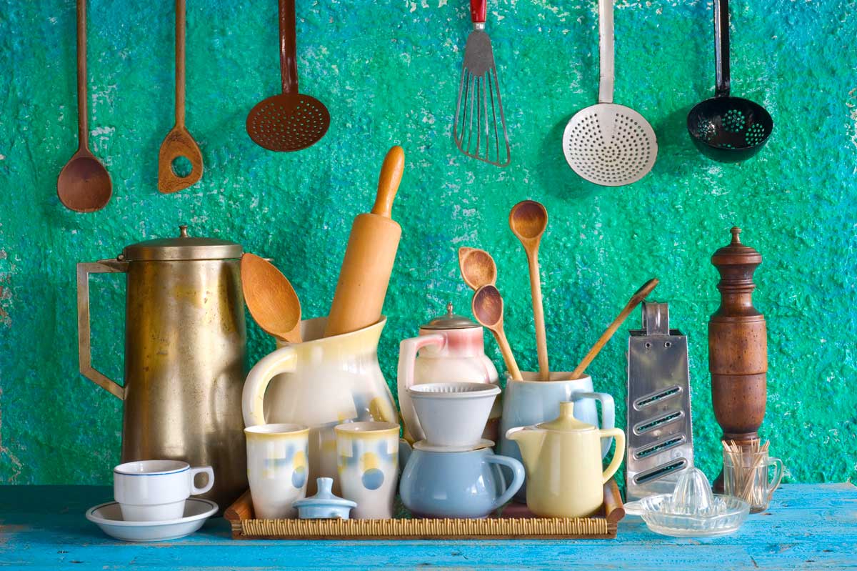 A collection of vintage kitchen utensils and crockery, including mugs, teapots, wooden spoons, and a rolling pin, arranged on a shelf in front of a turquoise textured wall with hanging ladles and spatulas.