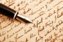 A close-up of a fountain pen resting on a piece of paper covered in elegant, cursive handwriting. The ink is a dark brown shade, and the writing appears to be part of an old-fashioned letter or document.