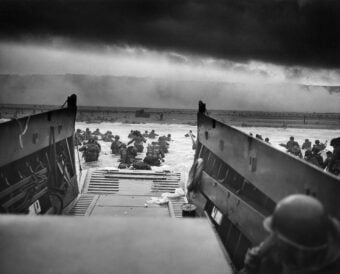 Black and white image of soldiers disembarking from a landing craft onto a beach during a military operation. They wade through shallow water towards the shore under an overcast sky, with distant smoke or haze visible.