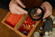 A person examines a gold ring with a magnifying glass above an open wooden box containing various rings and jewelry, with coins and other valuables scattered nearby on a wooden table.