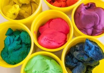 Top view of seven yellow cups each filled with colorful modeling clay in shades of yellow, red, purple, green, blue, light green, and pink, arranged closely together.