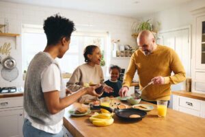 A family of four smiles and laughs together while making breakfast in a bright kitchen. Pancakes, fruit, and orange juice are on the counter as the parents cook and the children watch.