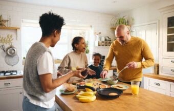 A family of four smiles and laughs together while making breakfast in a bright kitchen. Pancakes, fruit, and orange juice are on the counter as the parents cook and the children watch.