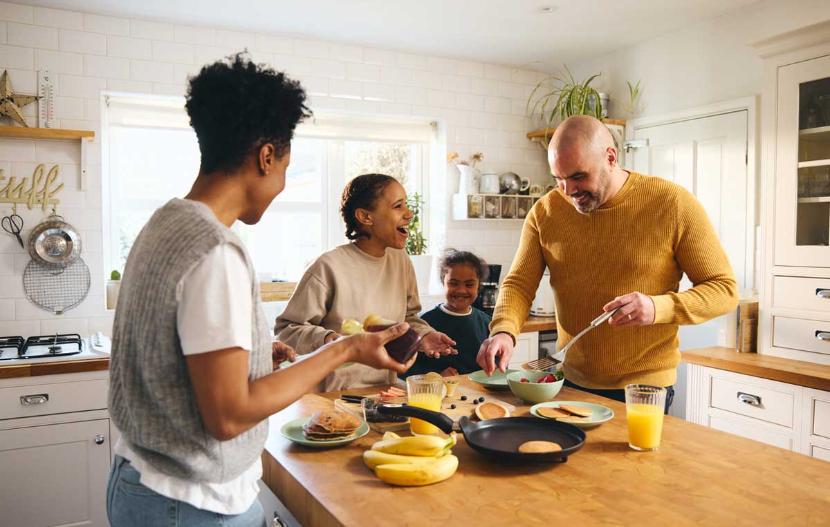 A family of four smiles and laughs together while making breakfast in a bright kitchen. Pancakes, fruit, and orange juice are on the counter as the parents cook and the children watch.