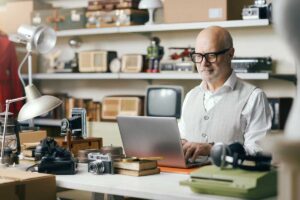 A man with glasses and a white vest types on a laptop at a desk cluttered with vintage cameras, books, and electronics, in a room filled with shelves of retro radios and collectibles.