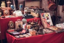 A table is covered with vintage items, including clocks, a typewriter, a rotary phone, decorative boxes, and colorful posters. The items are arranged on a red tablecloth, creating an eclectic display.