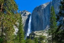 A tall waterfall cascades down a rugged cliff surrounded by pine trees and rocky terrain under a clear blue sky.