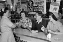 A waitress serves three smiling teenagers sitting at a diner counter. The teens have drinks, and a boy wears a sweater. Signs above advertise breakfast and hamburger specials. The scene appears to be from the 1950s.