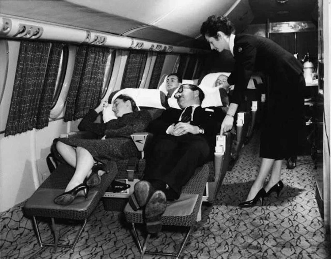 A black-and-white photo shows three airline passengers reclining and resting on spacious seats, while a flight attendant in a uniform stands beside them, assisting or conversing, inside a vintage airplane cabin.