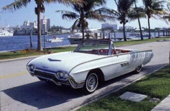 A vintage white convertible car is parked by a waterfront street lined with palm trees. Boats and buildings are visible across the water under a partly cloudy sky.