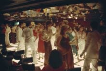 A crowded disco scene from the 1970s or 1980s with people dancing on a brightly lit floor, dressed in vintage clothing. A mirrored ceiling reflects the lively group, creating a vibrant and energetic atmosphere.