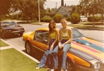 Two women in 1970s attire sit on the hood of a colorful, striped muscle car parked on a suburban street, with houses, trees, and another parked car visible in the background.