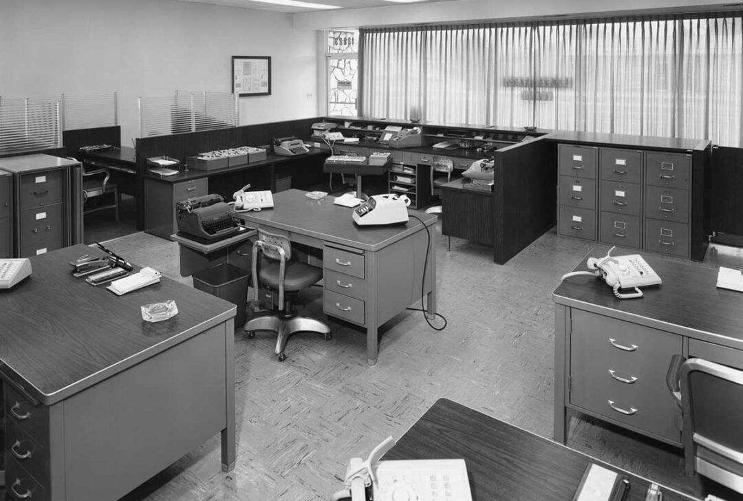 Black-and-white photo of a mid-20th century office with several desks, typewriters, rotary phones, filing cabinets, chairs, and paperwork, arranged in an organized, tidy layout.