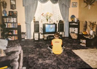A young boy sits on a carpeted floor watching TV in a living room with blue curtains, bookshelves, a stereo, VHS tapes, and floral decorations. The room has a cozy, vintage 1990s feel.