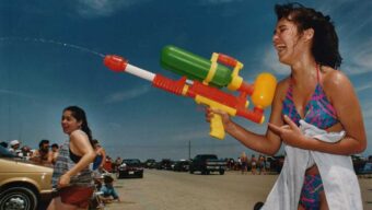 A young woman in a colorful swimsuit laughs while spraying a large water gun on a sunny beach, with cars and other people in the background.