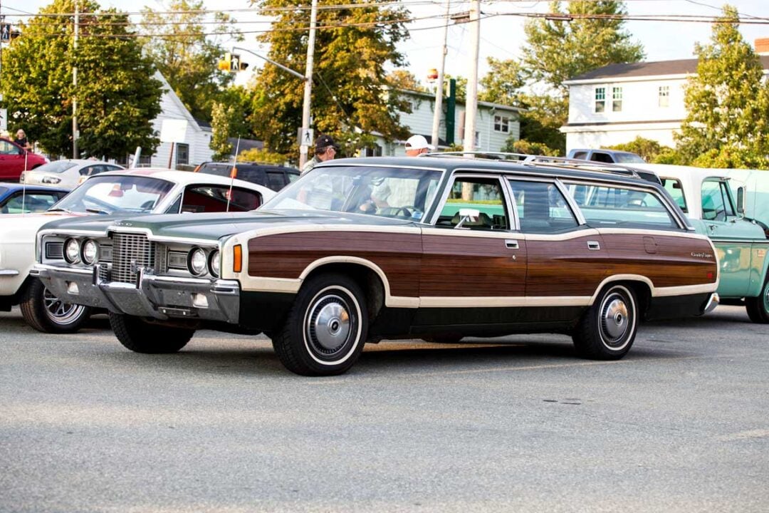 A vintage brown station wagon with wood paneling is parked at a classic car show, surrounded by other retro vehicles. Trees and houses are visible in the background.