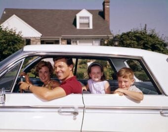 A smiling family of four sits in a white car parked in front of a suburban house; the father is driving, the mother sits next to him, and two children are in the back seat, all appearing happy.