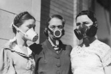 Three women in vintage clothing stand together outside, each wearing a different style of gas mask. The image is black and white, and the background is a brick or stone wall.