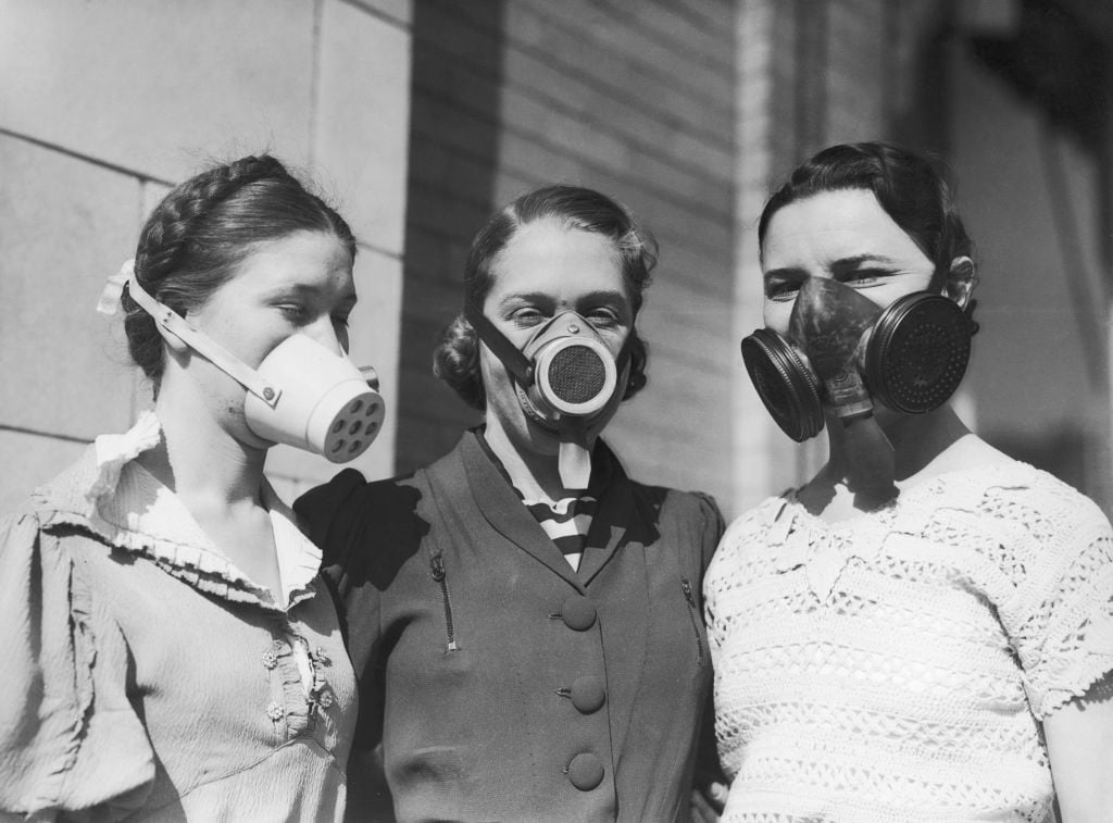Three women wearing vintage gas masks stand close together outside, dressed in early 20th-century clothing. The photo is black and white, with brick and stone walls visible in the background.