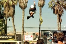 A person wearing a helmet is airborne on rollerblades, jumping high above a ramp with palm trees and spectators in the background on a sunny day.
