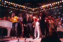 A large group of musicians sing together on a brightly-lit stage at the Live Aid concert, with a "Live Aid" logo visible on the backdrop and a crowd of enthusiastic performers in the background.