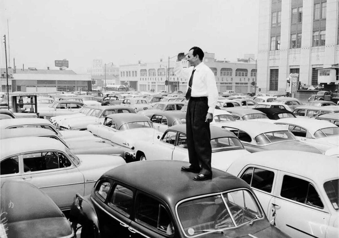 A man in a white shirt and tie stands on the roof of a car in a crowded parking lot, shading his eyes as he looks out over rows of parked vintage cars and city buildings in the background.