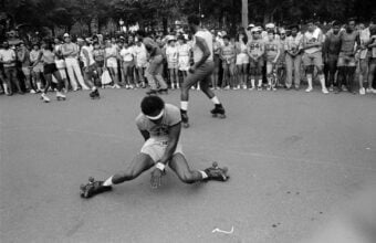 A person in athletic gear does a dramatic split on roller skates during a street performance, while a large crowd watches from the sidewalk in a park setting.