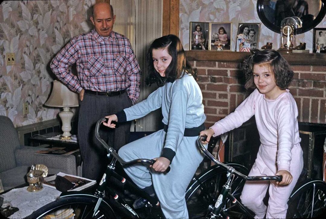 Two young girls in pajamas smile while sitting on bicycles indoors. An older man stands behind them, also smiling. The room has patterned wallpaper, a brick fireplace, and family photos on the mantel.