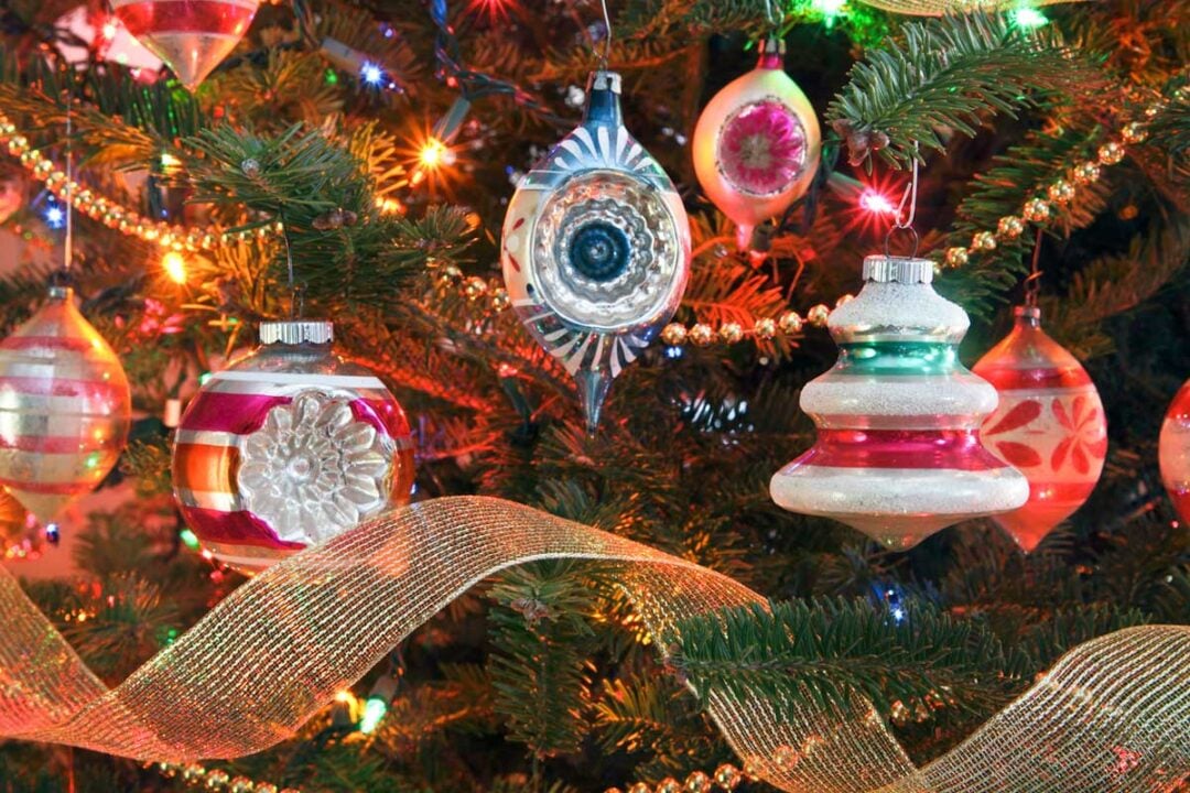 Close-up of a decorated Christmas tree with colorful vintage glass ornaments, gold beads, multi-colored string lights, and a sheer gold ribbon wrapped around the branches.
