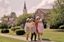 Four girls and one boy dressed in colorful, vintage 1960s-style clothing walk on a sidewalk past a suburban house with a church steeple in the background on a sunny day.