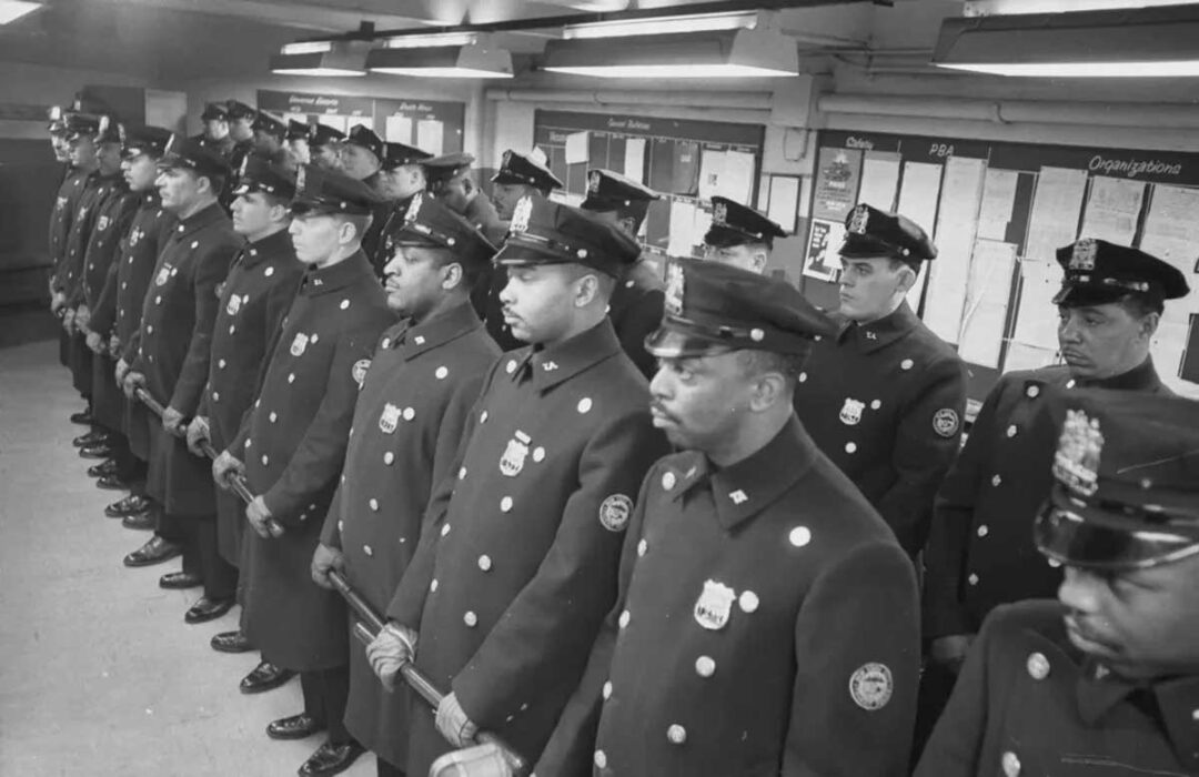 A group of uniformed police officers stands in a line indoors, facing forward. Notice boards and papers are visible on the wall behind them. The officers appear focused and attentive.