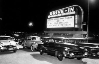 Black-and-white photo of classic cars parked at a drive-in theater at night, with a large sign showing films starring Gregory Peck, Anne Baxter, Mae West, and W.C. Fields.