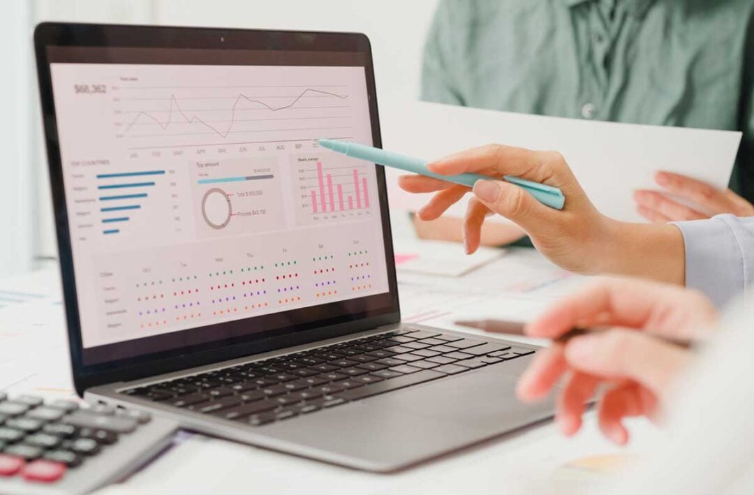 Two people review data visualizations on a laptop screen, including charts, graphs, and statistics. One person points at the screen with a pen while the other holds a paper document.