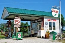 A vintage Sinclair gas station with a green roof, classic gas pumps, old signs, and a 15-cent gas price display, set on a gravel lot with blue sky and green trees in the background.