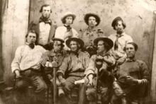 A vintage black-and-white photo shows eight men in 19th-century clothing posing for a group portrait. Some wear hats and hold objects, with a neutral studio backdrop behind them.
