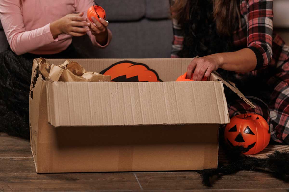 Two people sitting on the floor unpack Halloween decorations from a cardboard box, including small pumpkin-shaped buckets. Only their arms and torsos are visible.