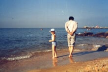 A child in a white hat and swimsuit stands on a sandy beach next to an adult, who is walking along the shore with hands behind their back. The sea is calm, and a few people are seen in the distance.