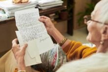 An elderly woman wearing glasses and a yellow sweater sits and reads a handwritten letter, holding the letter in both hands. A cozy blanket covers her lap, and a table with papers is visible in the background.