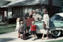 A family stands by a light blue car in a driveway. The father loads a suitcase into the trunk, while the mother and three young children, dressed in 1950s-style clothing, stand nearby in front of a suburban house.