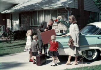 A family stands by a light blue car in a driveway. The father loads a suitcase into the trunk, while the mother and three young children, dressed in 1950s-style clothing, stand nearby in front of a suburban house.